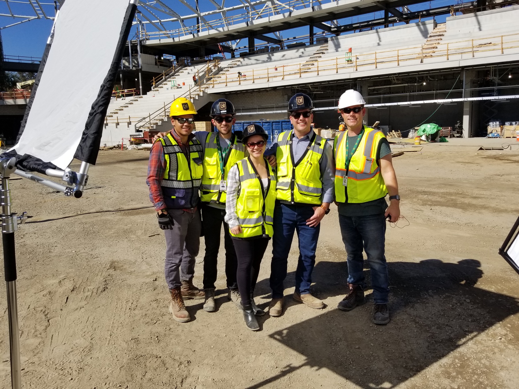 Five crew members in safety vests and hardhats with lighting equipment inside the unfinished LAFC stadium